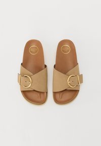 Des sandales à glissière à bandes croisées brunes avec un grand accent en boucle dorée, présentant une texture lisse et une semelle intérieure marron clair.
