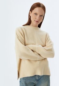 Beiger Wollpullover mit lockerer Passform, gerippten Manschetten und seitlichen Schlitzen. Er verfügt über ein dezentes weißes Stickdetail an der Vorderseite.