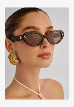 Vrouw met naar achteren gekamd haar die ovalen tortoise-shell zonnebrillen draagt, grote gouden spiraalvormige oorbellen en een witte geknoopte ketting.