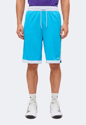 Homme portant un short de sport bleu vif avec bordure blanche et cordon, des chaussettes blanches de sport et des baskets blanches, debout devant un fond uni.