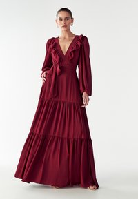 Burgundi ilgos maxi suknelė su volangų apykakle, ilgomis rankovėmis ir juosmeniu surišimu. Ypatumai: sluoksniuota sijonas ir plazdanti audinio tekstūra.