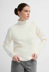 Sweter golf w kolorze kremowym o gładkiej fakturze, z opadającymi ramionami i długimi rękawami, zestawiony z szarymi spodniami. Minimalistyczny design.