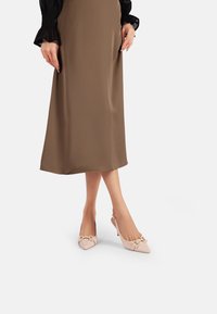 Tacchi slingback in pelle lucida rosa con punta affilata e dettagli dorati, abbinati a una lunga gonna in satin marrone.