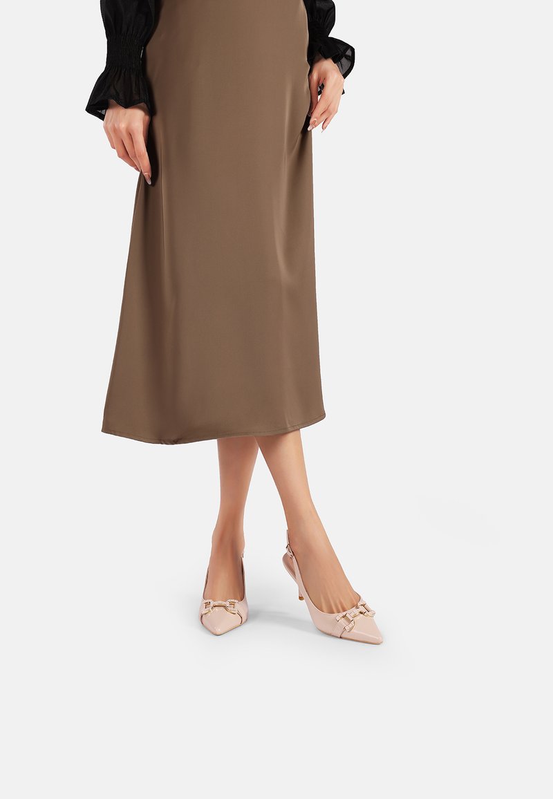 Tacchi slingback in pelle lucida rosa con punta affilata e dettagli dorati, abbinati a una lunga gonna in satin marrone.