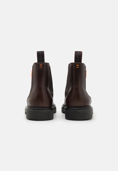 Brown leather Chelsea boots feature elastic side panels, pull tabs, and a thick black sole. The design includes orange accents on the side.