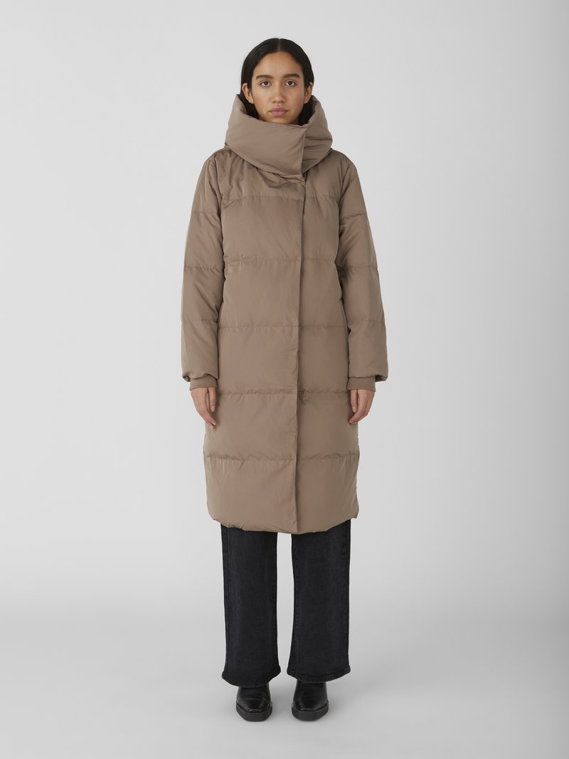 Manteau long beige en duvet avec col oversized, design matelassé rectangulaire et fermeture à boutons devant, associé à un jean noir à jambes larges et des chaussures noires.