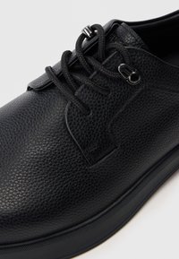 Chaussure en cuir noir avec une surface texturée, bout rond et semelle plate en caoutchouc. Équipée de lacets noirs et d'un accent en métal argenté.