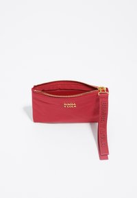 Bolsito rojo con cremallera dorada y detalle de logotipo, con una superficie texturizada y una correa de tela para la muñeca. Diseño compacto, adecuado para objetos pequeños.