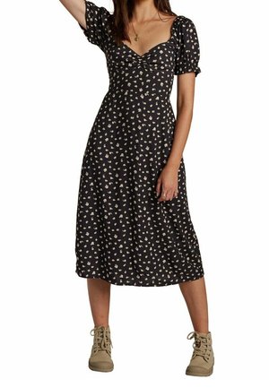 Femme portant une robe midi noire à petits imprimés floraux blancs, manches bouffantes, et bottines beiges, debout avec un bras levé.