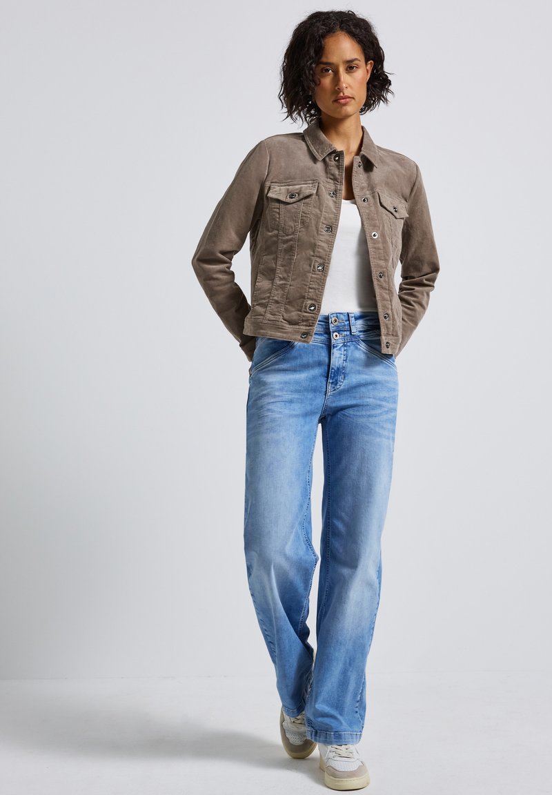 Brun kordfløyelsjakke med knappelukking, fire lommer, båret över en vit topp, i kombination med ljusblå vida jeans och beige sneakers.