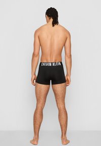 Schwarze Calvin Klein Boxer mit einer weißen Logo-Gummibund, die eine enge Passform und glatte Textur aufweisen, von hinten betrachtet.
