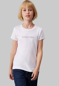 Camiseta blanca de manga corta hecha de algodón, con un logo gris de "Calvin Klein" centrado en el frente. Ajuste casual, cuello redondo.