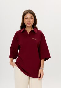 Polo shirt oversize color marrone con colletto, maniche corte e un sottile logo "MIRONS" in oro. Indossato con pantaloni di colore chiaro.