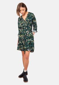 Blumenkleid in Grüntönen mit V-Ausschnitt, langen Ärmeln und Taillengürtel. Der Stoff hat ein strukturiertes Muster und wird mit schwarzen KnöchelStiefeln kombiniert.