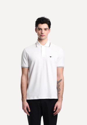 Giovane uomo con capelli corti e scuri che indossa una maglietta polo bianca con bordi neri, pantaloni neri e un tatuaggio sull'avambraccio sinistro.