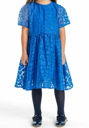 Bambina che indossa un vestito blu a fantasia fino al ginocchio con maniche corte trasparenti, calze scure e scarpe Mary Jane blu scuro.