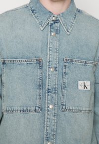 Camicia in denim chiaro con chiusura frontale con bottoni, due tasche sul petto e una toppa con logo CK sulla tasca sinistra. Caratterizzata da un colletto appuntito.