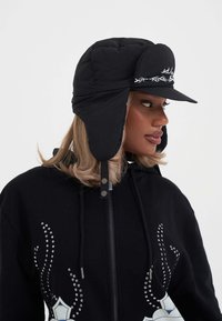 Cappello imbottito nero con alette per le orecchie e una visiera curva. Presenta ricami bianchi sulla parte anteriore e una superficie liscia e lucida.