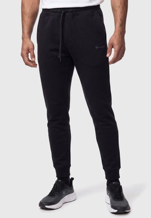 Hombre con pantalones de jogging negros con cordón y zapatos deportivos en blanco y negro sobre un fondo claro y liso.