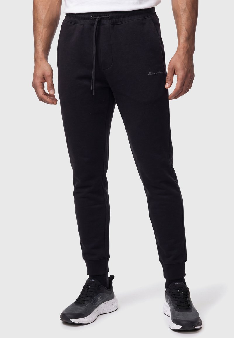 Homme portant un pantalon de jogging noir à cordon et des chaussures de sport noires et blanches sur un fond uni clair.