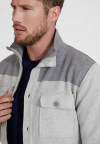 Graue und hellgraue Kollerjacke mit Reißverschluss, ausgestattet mit zwei Klappentaschen und einer weichen Textur. Getragen über einem marineblauen Hemd.