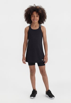 Girl with curly hair wearing a black sleeveless dress over black shorts and black sneakers, standing against a plain white background.