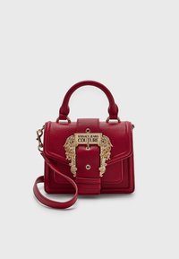 Punane kivitooted nahast käekott, millel on ülemine käepide, eemaldatav rihm ja suur kuldne ornamenteeritud pandla, millel on graveering "Versace Jeans Couture."