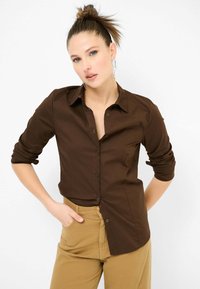 Camicia marrone con bottoni, realizzata in tessuto liscio, con colletto a punta e maniche arrotolate, abbinata a pantaloni khaki. Design semplice, forma aderente.
