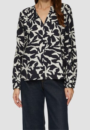 Kvinde iført en sort- og hvid bluse med blomsterprint, lange ærmer med elastiske manchetter og mørkeblå denimjeans.