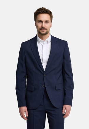 Mężczyzna z krótkimi brązowymi włosami i brodą, ubrany w granatowy garnitur i białą koszulę, stoi na tle jednolitego jasnego tła.