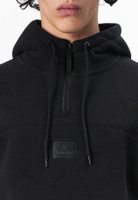 Primo piano di una persona che indossa un hoodie in felpa nera con zip frontale, coulisse e patch del logo Timberland sul petto.