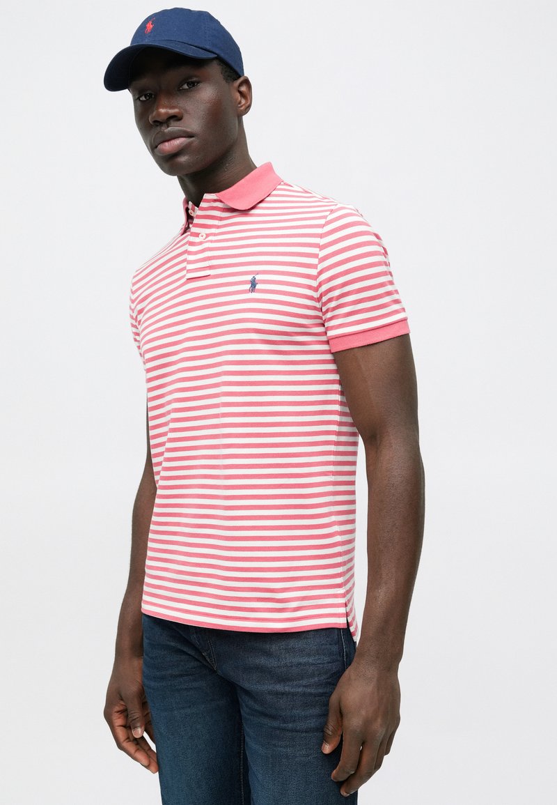 Red and white striped polo shirt with a navy collar, featuring a small logo on the chest. Worn with dark blue jeans and a matching cap.