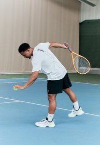 Tennisspiller i en hvit skjorte og svarte korte bukser gjør seg klar til å serve en grønn tenniskule. Han bruker et lyseoransje racket og har på seg hvite joggesko.