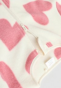 Vêtement en polaire crème avec de grands motifs de cœurs roses, doté d'une fermeture éclair à l'avant et d'une texture douce. Possède une étiquette indiquant "Next Baby."