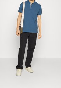 Mies, joka on pukeutunut siniseen polo-paitaan, mustiin housuihin ja valkoisiin tennareihin, kantaa vihreää ja beigeä olkalaukkua yksinkertaista taustaa vasten.