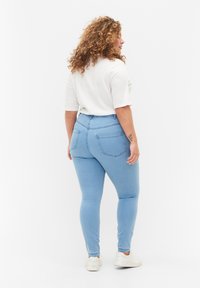 Jeans ajustados de color azul claro con cintura alta, cuentan con dos bolsillos traseros y una textura de denim suave. Combinados con una camiseta blanca y zapatillas.