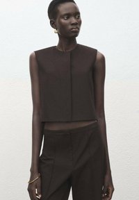 Mujer con cabello corto, vestida con top sin mangas marrón oscuro y pantalones a juego, accesorios de pendientes de aro dorados y pulsera, fondo neutro.