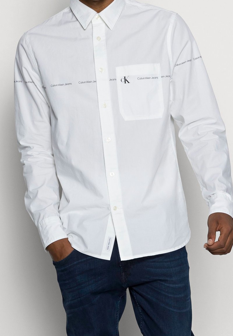Vit långärmad skjorta med knappar i bomull, med en bröstficka och tryckt varumärke längs ärmarna i svart.