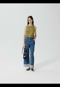 Junge Frau trägt ein olivfarbenes ärmelloses Oberteil, weite blaue Jeans mit Umschlag und schwarze Ballerinas, hält eine kleine marineblaue Handtasche mit roten und orangen Details.