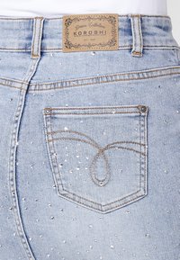 Dos de jean denim bleu clair avec coutures marron, petits clous argentés et une étiquette rectangulaire marron de la marque Koroshi sur la ceinture.