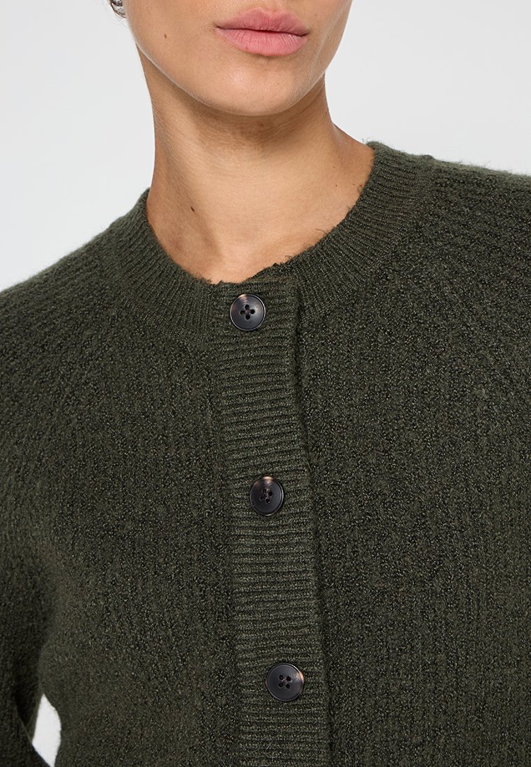 Dunkelgrüner gestrickter Cardigan mit Rundkragen, strukturiertem Stoff und vier Knopfverschlüssen vorne. Die Knöpfe sind schwarz und rund.