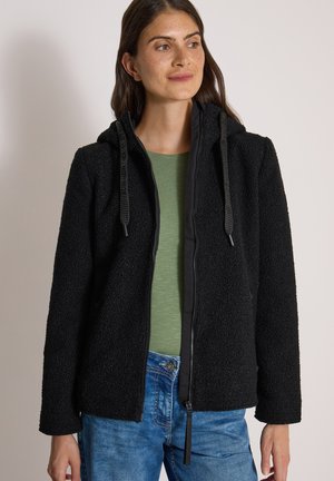 Femme portant une veste à capuche noire zippée par-dessus une chemise verte et un jean bleu, regardant légèrement vers sa droite avec une expression neutre.