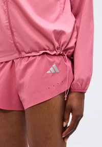 Κοντινό πλάνο ενός ατόμου που φορά ροζ σορτς Adidas και αντίστοιχο μπουφάν με ρυθμιζόμενο κορδόνι στη μέση, εστιάζοντας στον κάτω κορμό και το δεξί χέρι.