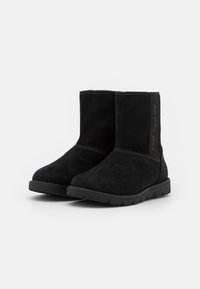 Bottines en suede noir avec une semelle en caoutchouc, présentant une finition texturée et un détail de logo latéral. Coupe droite, design minimaliste.