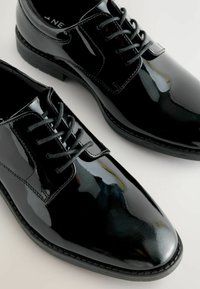Chaussures habillées en cuir verni noir avec un bout pointu, fermeture à lacets et surface texturée, présentant une finition lisse et une semelle solide.