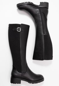 Un paio di stivali in pelle neri alti fino al ginocchio, con pannelli in tessuto, zip laterale, fibbia decorativa e tacco basso chunky, su sfondo bianco.