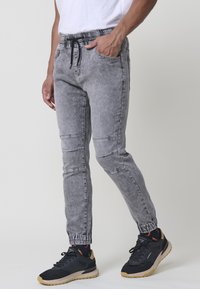 Pantalones de chándal grises de mezclilla con cintura elástica y cordón, que cuentan con bolsillos laterales y puños acanalados. Combinados con zapatillas negras.
