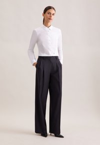 Kobieta stojąca z rękami w kieszeniach, ubrana w białą koszulę zapinaną na guziki, szerokie czarne spodnie i czarne szpilki z ostrym noskiem na jednolitym tle.