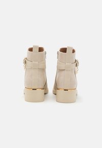 Bottines beiges à finition texturée, avec des zips latéraux, des accents dorés et un talon carré. Équipées de deux sangles à l'arrière.