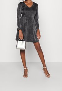 Robe noire scintillante avec un col en V, des manches longues et une jupe évasée. Associée à un sac à main argenté et des talons transparents à lanières.